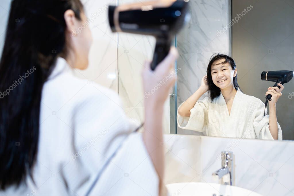 Happy asian child girl drying hair by blowing warm air with hair dryer and looking at the mirror in bathroom,beautiful smiling female in bathrobe using a hairdryer after taking a shower dries wet hair