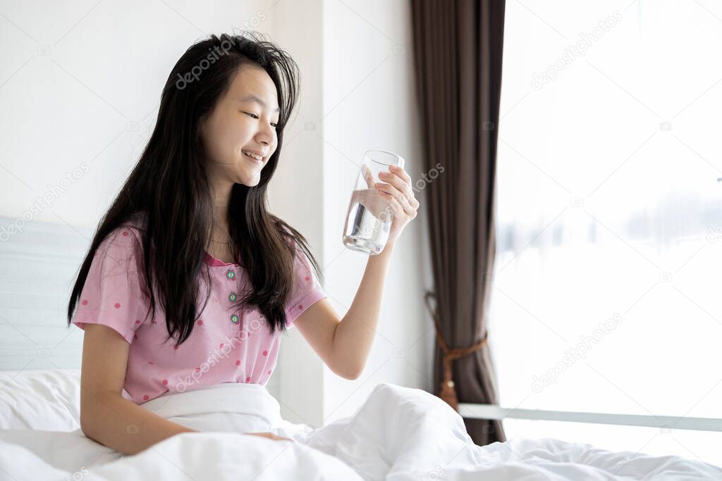 Smiling asian young woman holding a glass of water while sitting in bed after waking up in the morning,thirsty girl enjoy drinking fresh water to refresh,body healthy and skin health care concept