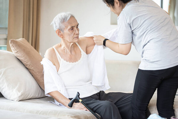 Asian female caregiver taking care of helping elderly patient get dressed,wearing clothes or undressing for senior mother,depressed old woman with amnesia,loss of memory,daily life in a nursing home
