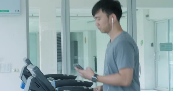 Junger asiatischer Mann läuft mit Kopfhörern auf dem Laufband im Fitnessstudio. — Stockvideo