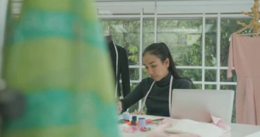 DS Ms Beautiful Asyalı kadın moda tasarımcısı skeçler modern Studio bir laptop arıyorum, notlar alarak masasında oturan.