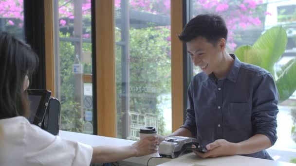 Красивая азиатка делает кофе на вынос для клиента, который платит с помощью бесконтактного мобильного телефона к системе кредитных карт — стоковое видео