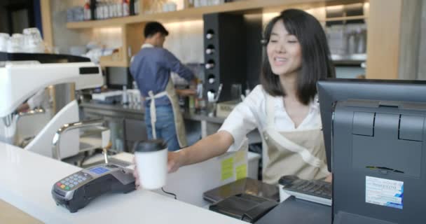 Bella donna asiatica rende caffè da asporto per un cliente che paga tramite telefono cellulare senza contatto al sistema di carte di credito — Video Stock