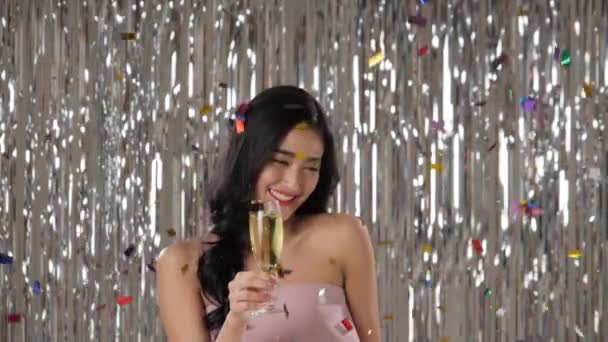 Beautiful asian woman holding glass of champagne dancing among confetti. — Stock Video