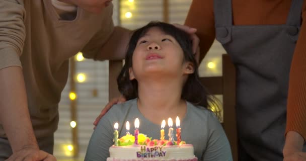 Um bonito menina asiática sentada à mesa e soprando velas no bolo de aniversário, enquanto sua família de pé atrás e cantar uma música para ela . — Vídeo de Stock