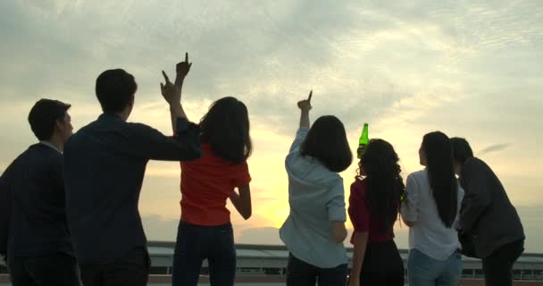 Grupo de jóvenes amigos bailando y divirtiéndose celebran con tostadas y tintineo levantando vasos en la fiesta de verano en la azotea con puesta de sol en el fondo . — Vídeo de stock