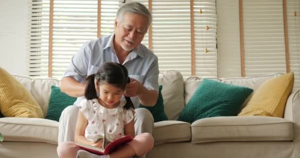 Asiático mayor abuelo atando pequeña niña cabello mientras ella leyendo libro en casa . — Vídeo de stock