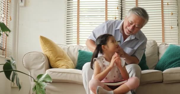 Asiático mayor abuelo atando pequeña niña cabello mientras ella leyendo libro en casa . — Vídeo de stock