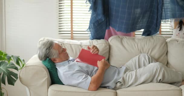 Senior schläft mit Buch auf dem Sofa im Wohnzimmer und seine Frau legt ihm Decke über. — Stockvideo