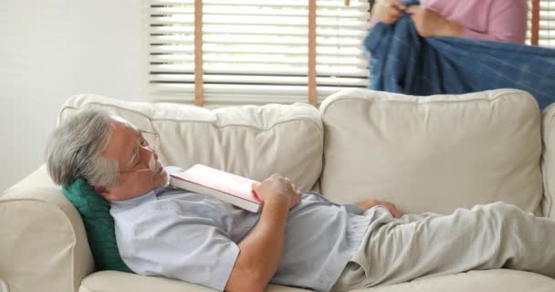 Senior schläft mit Buch auf dem Sofa im Wohnzimmer und seine Frau legt ihm Decke über. — Stockvideo