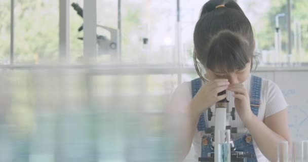 Pequeña estudiante asiática mira atentamente en el microscopio en la mesa, Niña haciendo experimentos científicos . — Vídeo de stock