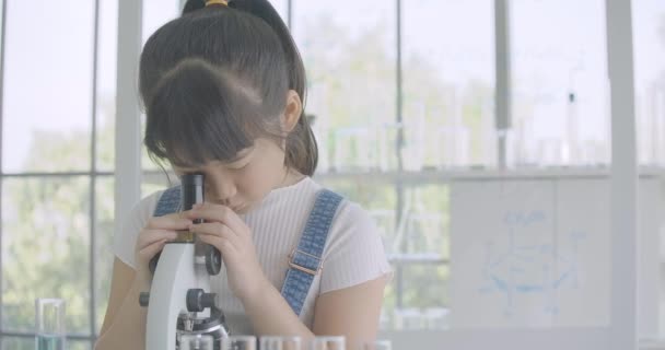Ασιάτισσα φοιτήτρια κοριτσάκι προσεκτικά φαίνεται σε μικροσκόπιο στο τραπέζι, κάνοντας πειράματα επιστήμης το μικρό κορίτσι. — Αρχείο Βίντεο