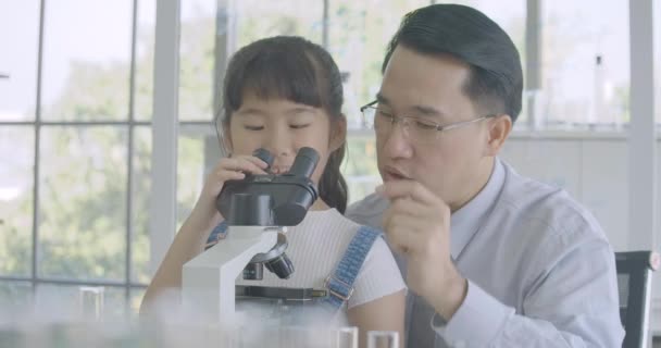 Asian male scientist teaching little girl scientific experiment using microscope in classroom laboratory. — Stockvideo