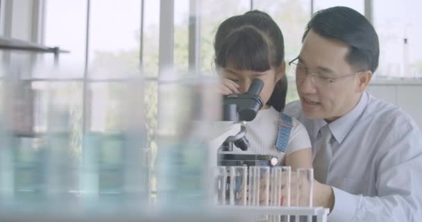 Asian male scientist teaching little girl scientific experiment using microscope in classroom laboratory. — Stock Video