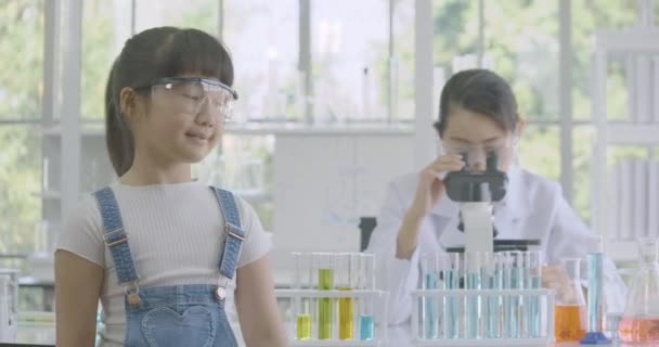 Porträt eines kleinen asiatischen Mädchens, das in einem Chemielabor in die Kamera lächelt, im Hintergrund arbeitet eine Wissenschaftlerin — Stockvideo