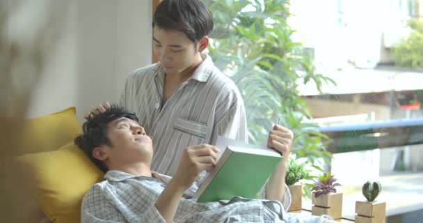 Unga asiatiska gay par sitta på fönsterbrädan och läsa bok hemma på morgonen — Stockvideo