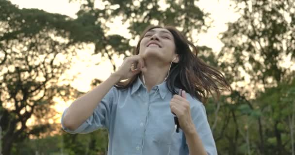 Young attractive asian woman listens to music with phone and headphones enjoying sound dances in a summer park. — Stock Video