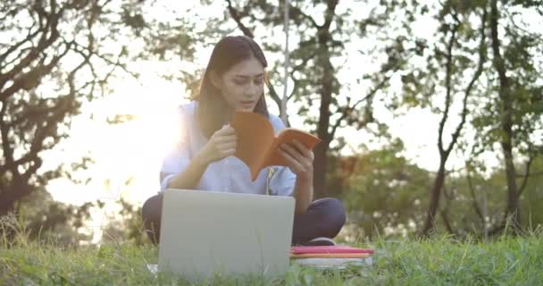 Jeune femme asiatique attrayant assis dans un parc d'été sur l'herbe et la lecture d'un livre au coucher du soleil . — Video