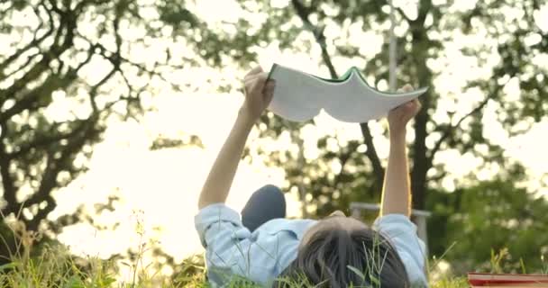 Young attractive asian woman reading book lying down on green grass in the park at sunset. — Stock Video