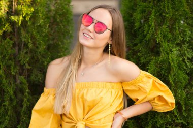 Parlak sarı bluz ile sarışın saçlı güzel genç kadın portresi açık omuz ve doğal yeşil zemin üzerine kırmızı güneş gözlüğü. Bir kız portresi yaz