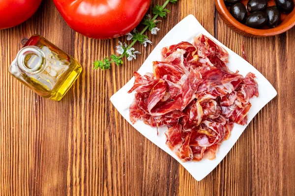 İştah açıcı bir plaka üzerinde İber Jambon dilimleri. Ahşap arka plan üzerinde. Zeytin yağı, taze domates, zeytin ve fesleğen içerir. Bir makale haute mutfağının ve ham tuzlama ve doğal curin bir işlemi boyunca gastronomik lüks olur ham et