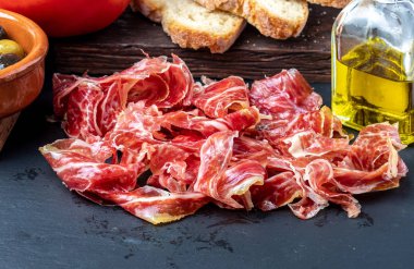 Ön planda iştah açıcı dilimler İber jambonu. Zeytinyağı, ekmek, taze domates, zeytin. Siyah arka plan. Rustik ve ev yapımı görünüm.