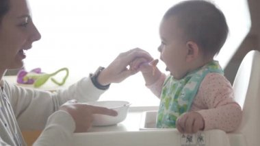 Genç mutlu anne bebeğine kaşıktan, bebek mamasından kendi başına yemeyi öğretiyor.