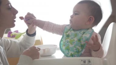 Genç mutlu anne bebeğine kaşıktan, bebek mamasından kendi başına yemeyi öğretiyor.