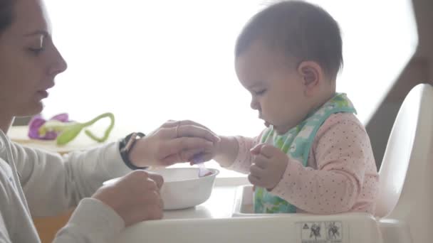Молодая счастливая мать учит своего малыша есть самостоятельно с ложки, детское питание — стоковое видео