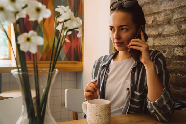 Jovem mulher bonita sentada em um café e falando ao telefone. A menina bebe café quente — Fotografia de Stock