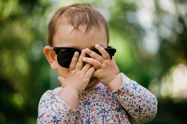 Малышка, играющая в пип-девочку, прячущую лицо. Летний фон. Солнечные очки — стоковое фото