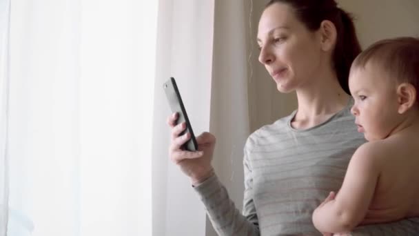 Mãe e bebê brincando com um telefone inteligente newr a janela na sala de estar em casa 4k — Vídeo de Stock
