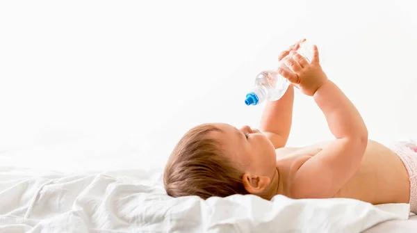 Дитячий малюк сидить на білому ліжку, посміхається і п'є воду з пластикової пляшки. Копіювати простір — стокове фото