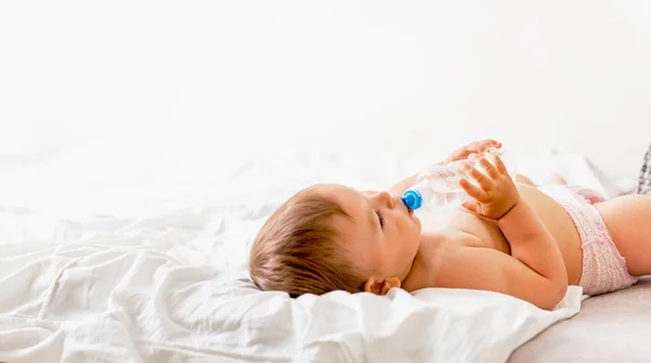 Kleinkind sitzt auf dem weißen Bett, lächelt und trinkt Wasser aus Plastikflasche. Kopierraum — Stockfoto