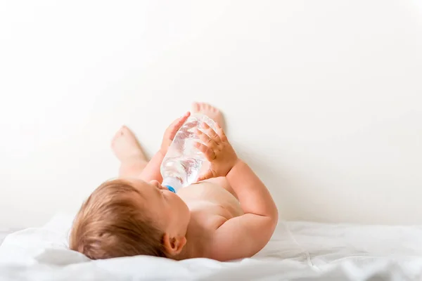 Kleinkind sitzt auf dem weißen Bett, lächelt und trinkt Wasser aus Plastikflasche. Kopierraum — Stockfoto