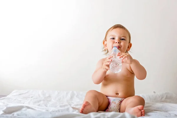 Bebê criança senta-se na cama branca, sorri e bebe água de garrafa de plástico. Espaço de cópia — Fotografia de Stock