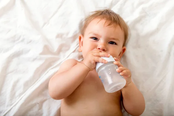 Дитячий малюк лежить на білому ліжку, посміхається і п'є воду з пластикової пляшки, копіює простір — стокове фото