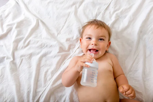 Baby liegt auf dem weißen Bett, lächelt und trinkt Wasser aus Plastikflasche, Kopierraum — Stockfoto