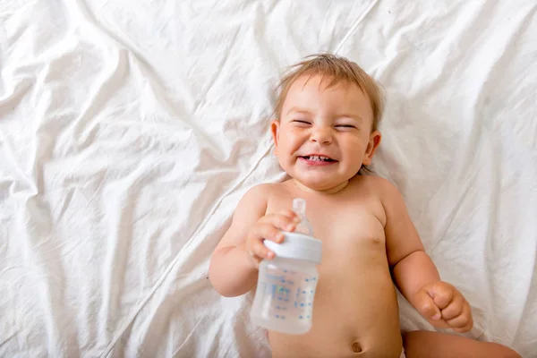 Дитячий малюк лежить на білому ліжку, посміхається і п'є воду з пластикової пляшки, копіює простір — стокове фото