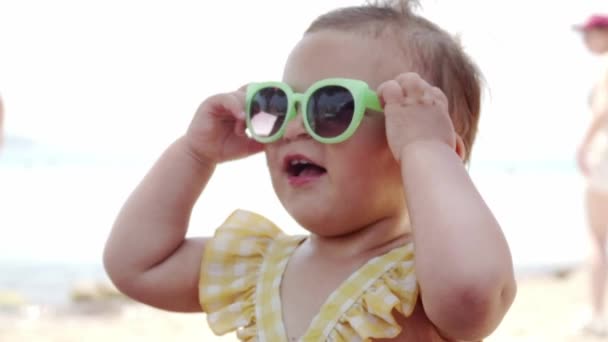 Ένα κοριτσάκι με γυαλιά ηλίου στην παραλία στο θέρετρο. Αστείο νήπιο. Να ταξιδεύεις με ένα μωρό. Καλοκαιρινή παραλία — Αρχείο Βίντεο