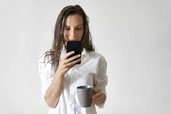 Menina bonita com cabelo molhado e xícara de mensagens de texto café no smartphone. Espaço cópia rotina da manhã — Fotografia de Stock