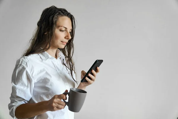 Menina bonita com cabelo molhado e xícara de mensagens de texto café no smartphone. Espaço cópia rotina da manhã — Fotografia de Stock