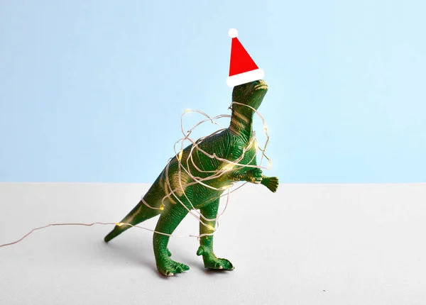 Divertido tiranosaurio verde Rex en sombrero de santa roja y luz de Navidad. Concepto mínimo azul — Foto de Stock