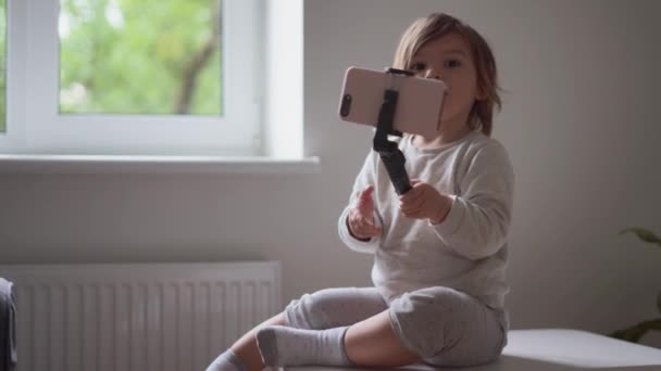 Küçük kız evde oturuyor ve selfie telefon çubuğu kullanıyor. Çocuklar için nüfuz ve sosyal medya — Stok video