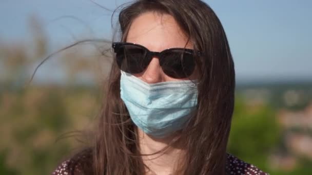 夏の日に屋外で顔の医療マスクとサングラスを持つ若い女性。新常態 — ストック動画