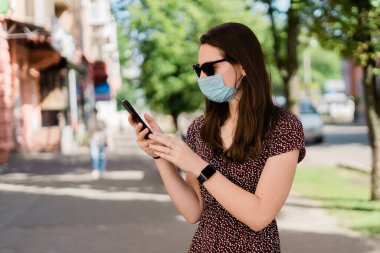 Tıbbi maskeli ve telefonlu genç bir kadın. Yaz açık havada yeni normal yaşam tarzı