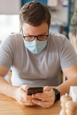 Kafkasyalı bir adam restoranda oturuyor ve tıbbi maske takıp telefon kullanıyor. Yeni Normal kavramı