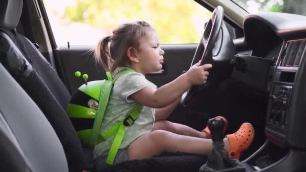 赤ん坊の幼児の女の子はステアリングホイールと車のフロントシートに座っている。学校に戻る — ストック動画
