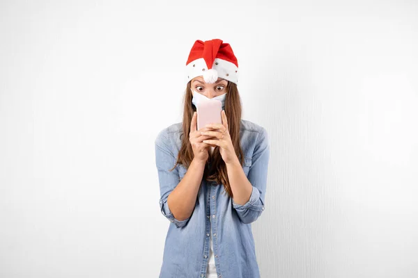 年轻的女人带着电话医疗面具和圣塔帽。圣诞网上购物检疫. — 图库照片