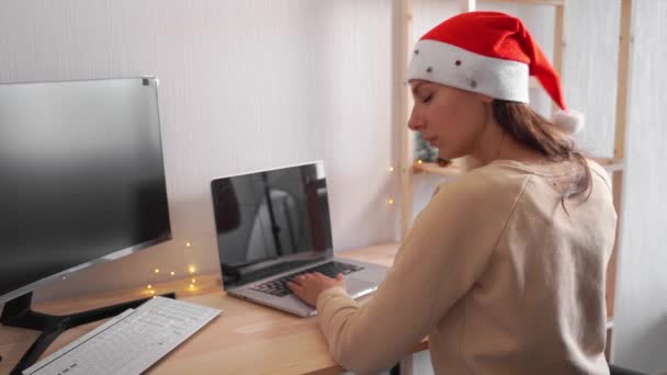 Mujer joven con sombrero de santa y luces de Navidad solo en la oficina. Compras en línea. Celebración de cuarentena — Vídeo de stock